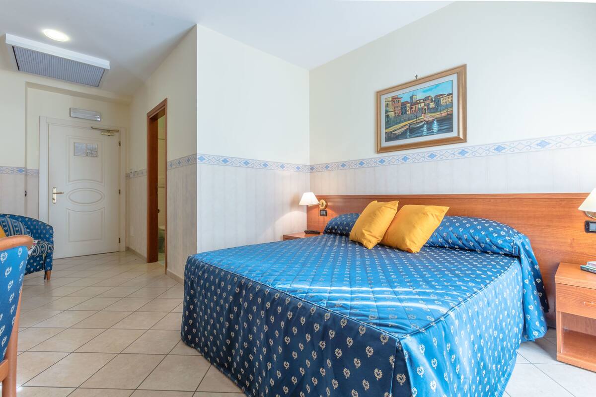 A hotel room with a large bed, a wooden headboard, a nightstand, a lamp, a framed picture on the wall, and a door leading to another room.