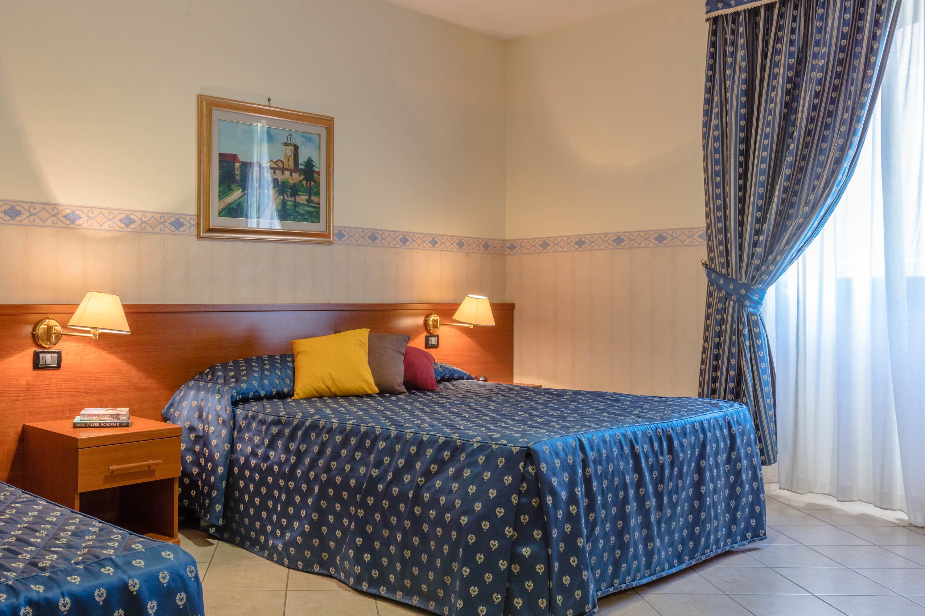 Una habitación de hotel con una cama, dos mesitas de noche, una lámpara, un cuadro en la pared y una ventana con cortinas.