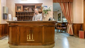 Un mostrador de recepción con detalles de madera y un gran emblema 'HT'. Un hombre está de pie detrás del mostrador, hay una planta y una cortina a la derecha.
