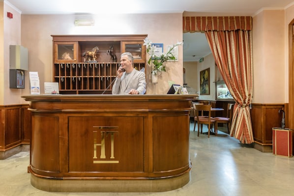 A reception desk with a wooden finish and a large 'HT' emblem. There is a man standing behind the desk, a plant, and a curtain to the right.