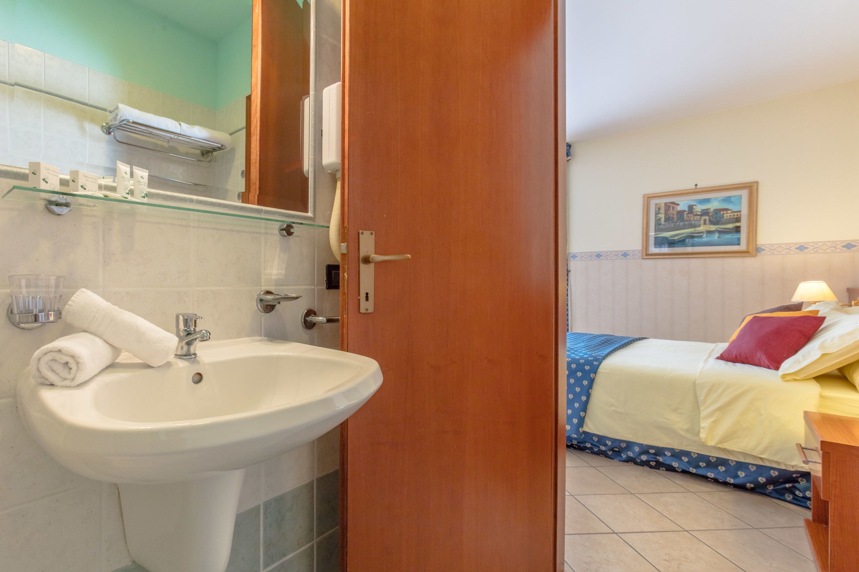 Une salle de bain équipée d’un lavabo, d’un miroir et d’un porte-serviettes. La pièce comprend un lit avec une couvre-lit bleue et un tableau accroché au mur.