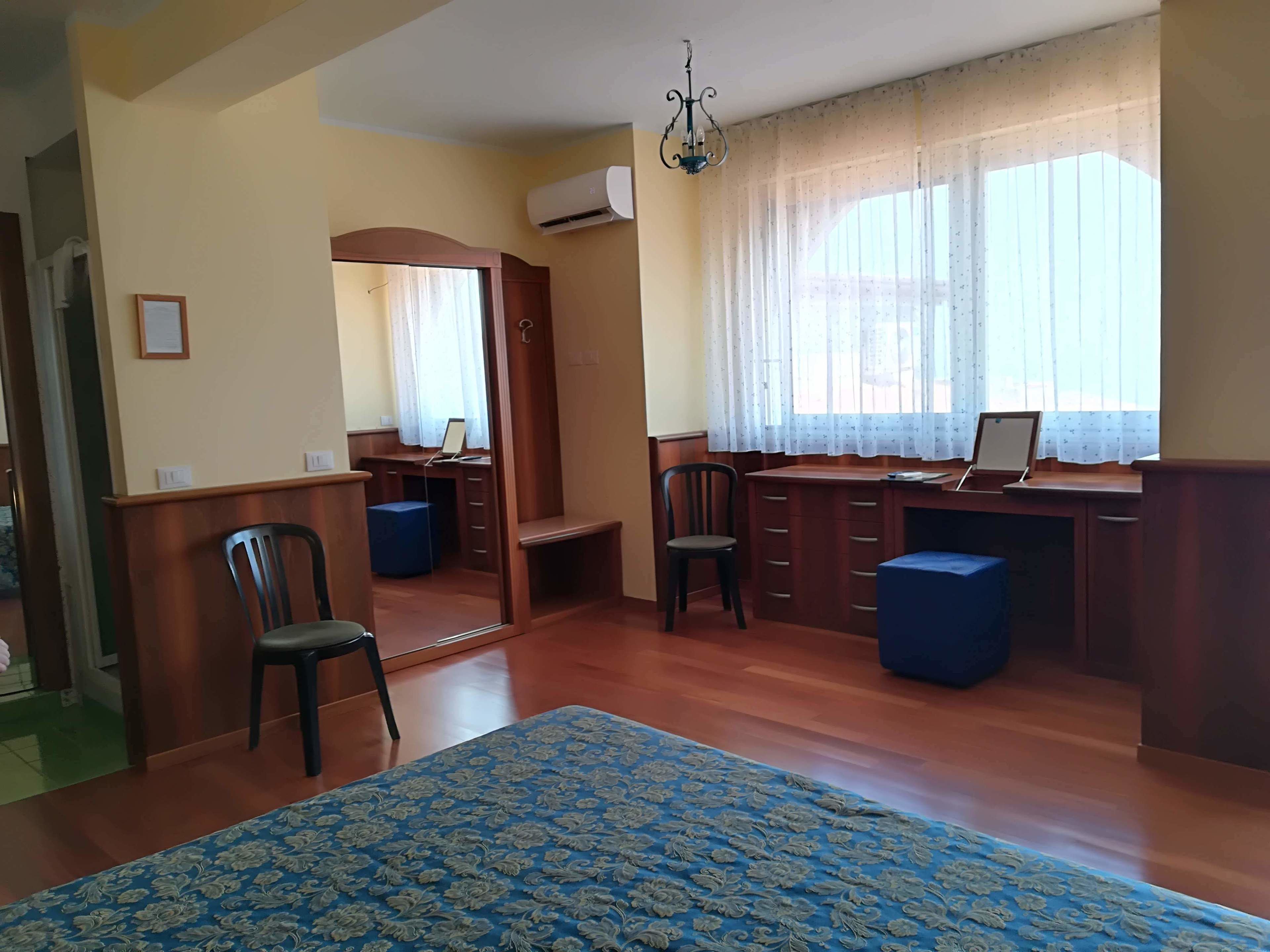 A room with a wooden desk, two chairs, a bed with a floral bedspread, a chandelier, and a window with sheer curtains.