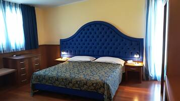 Quarto com cabeceira azul, colcha floral, mobiliário de madeira e janela com cortinas.