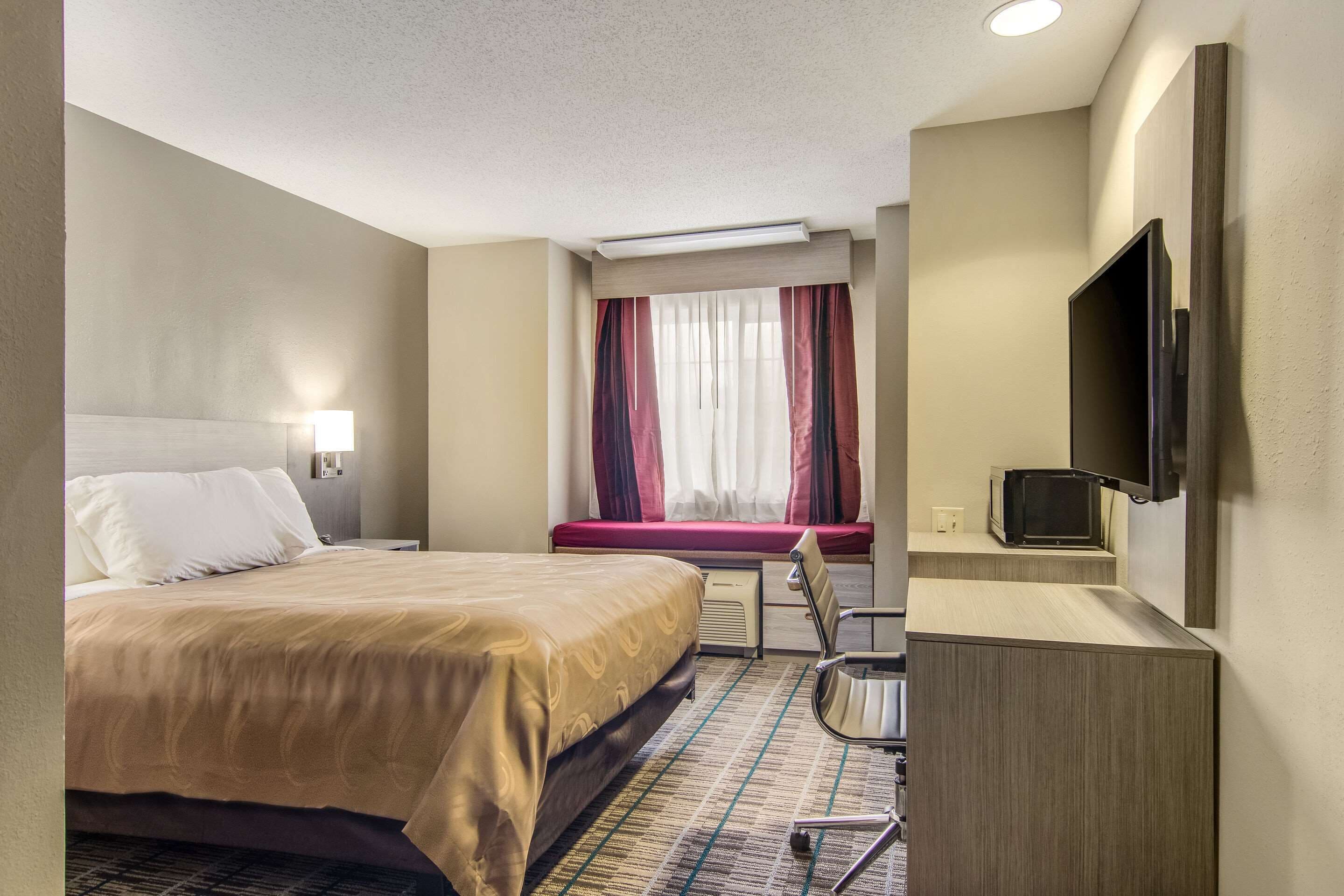 Una habitación de hotel con cama, escritorio, silla, televisión y ventana con cortinas.