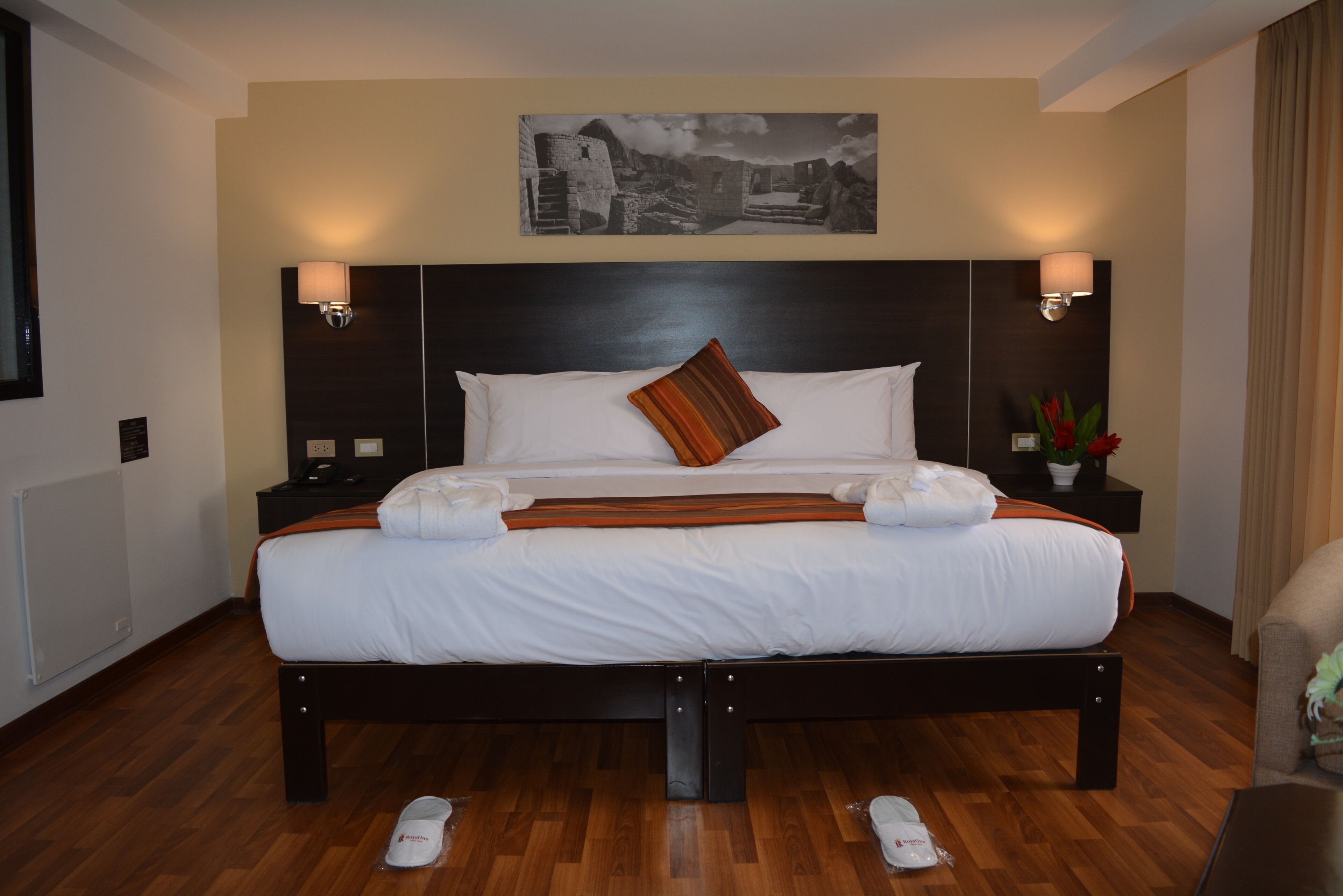 Una cama bien hecha con sábanas blancas y dos almohadas, con mesitas de noche y lámparas a ambos lados, en una habitación con suelo de madera y un cuadro en la pared.