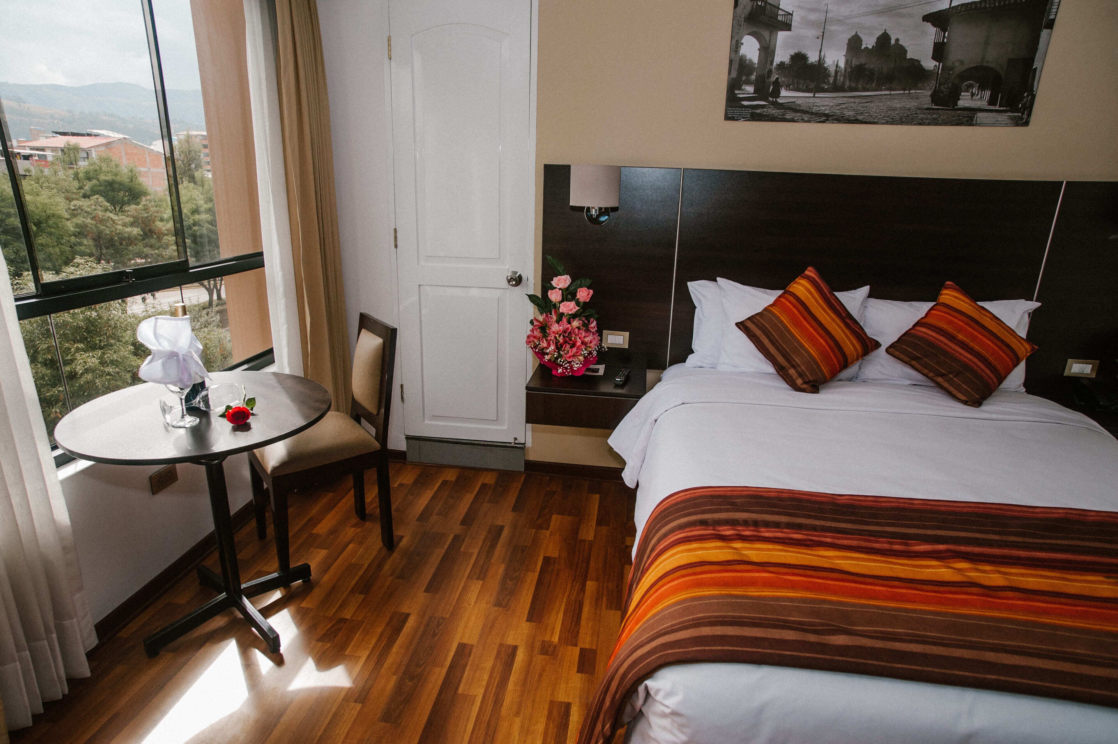 A hotel room with a bed, a small table with a chair, a nightstand with a lamp, and a view of the outside.
