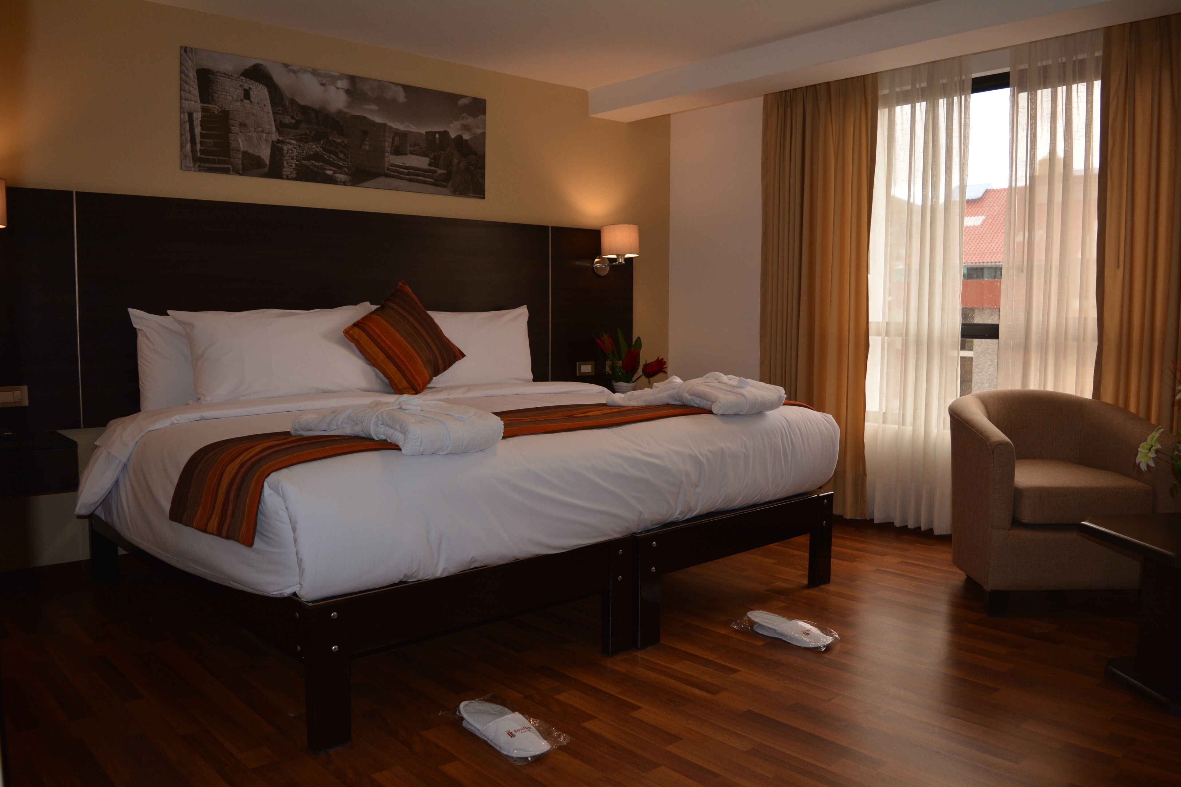 A hotel room with a large bed, a nightstand, a lamp, a chair, and a window with curtains.