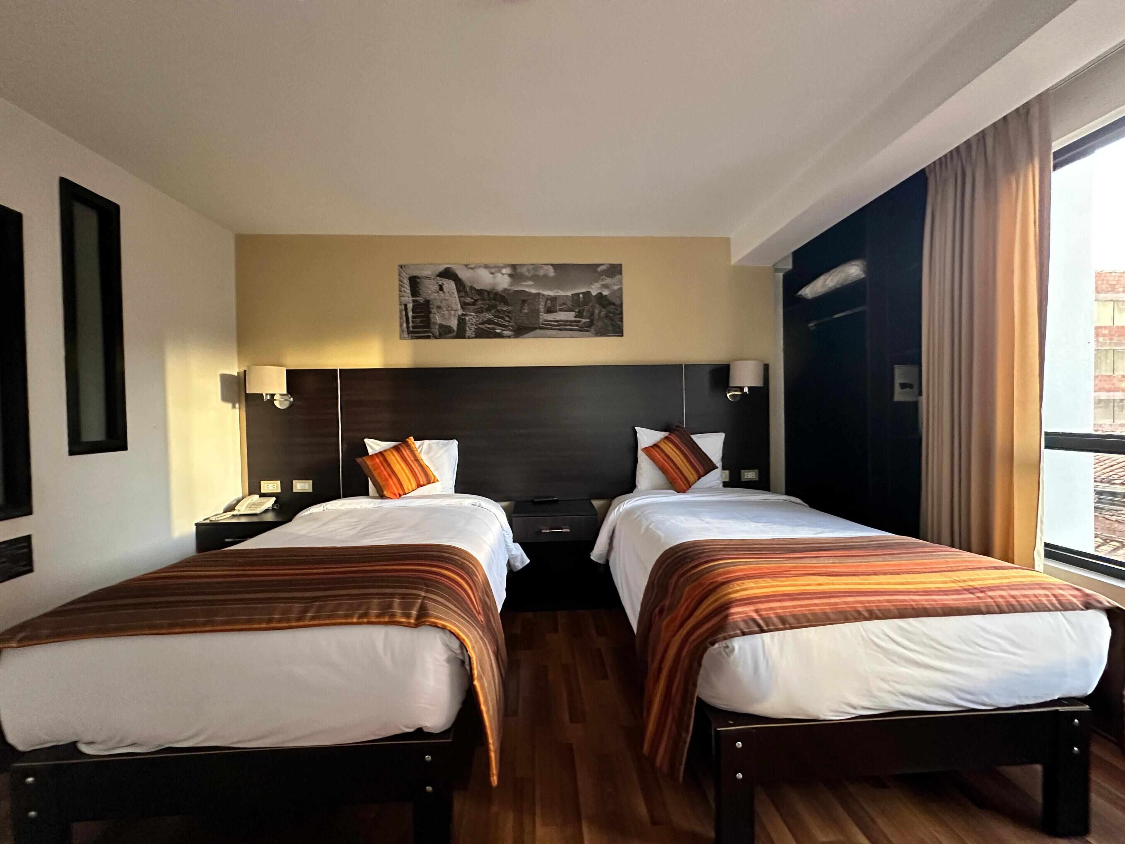 A hotel room with two beds, a nightstand with a lamp, a painting on the wall, and a window with curtains.