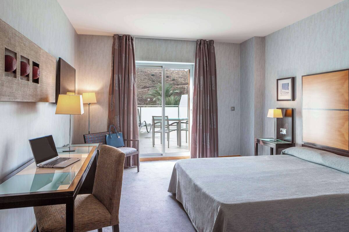 Habitación de hotel con cama, escritorio con una computadora portátil, una silla, mesa con una silla, balcón con mesa y sillas, y ventana con cortinas.