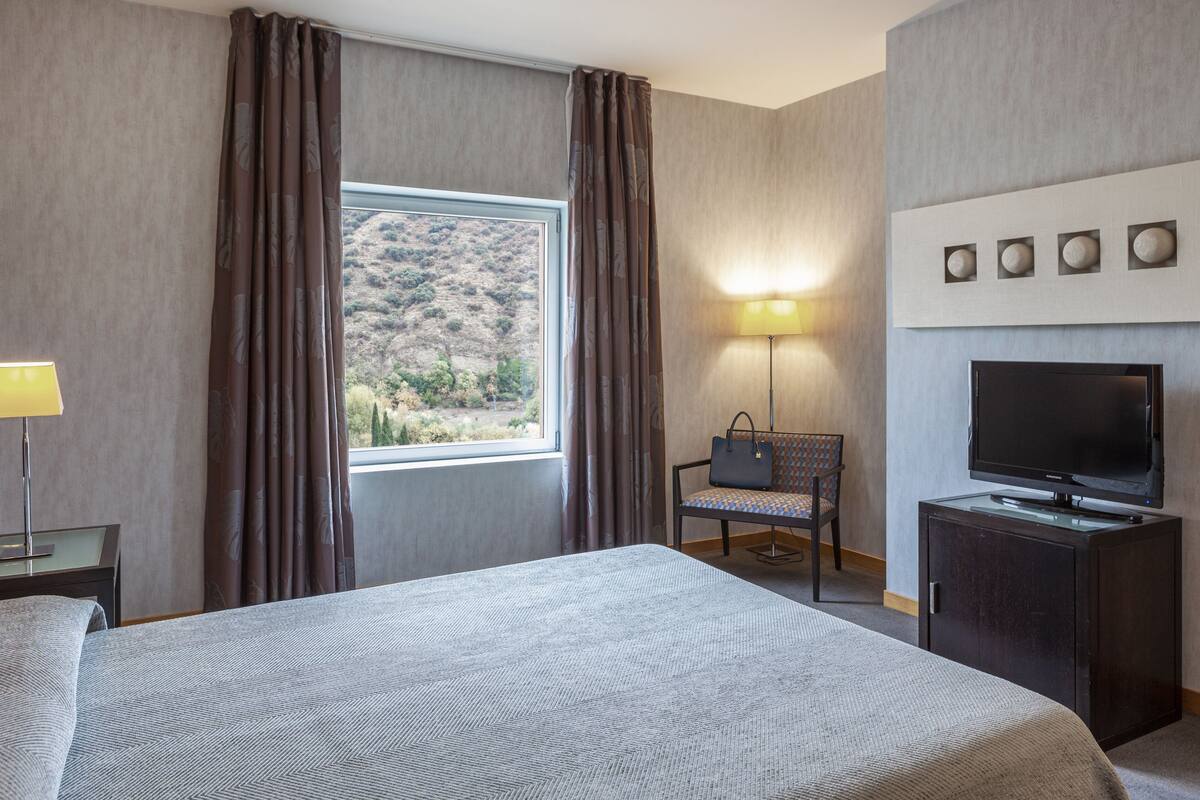 Una habitación de hotel con una cama grande, televisión, escritorio con lámpara, una silla y ventana con cortinas.