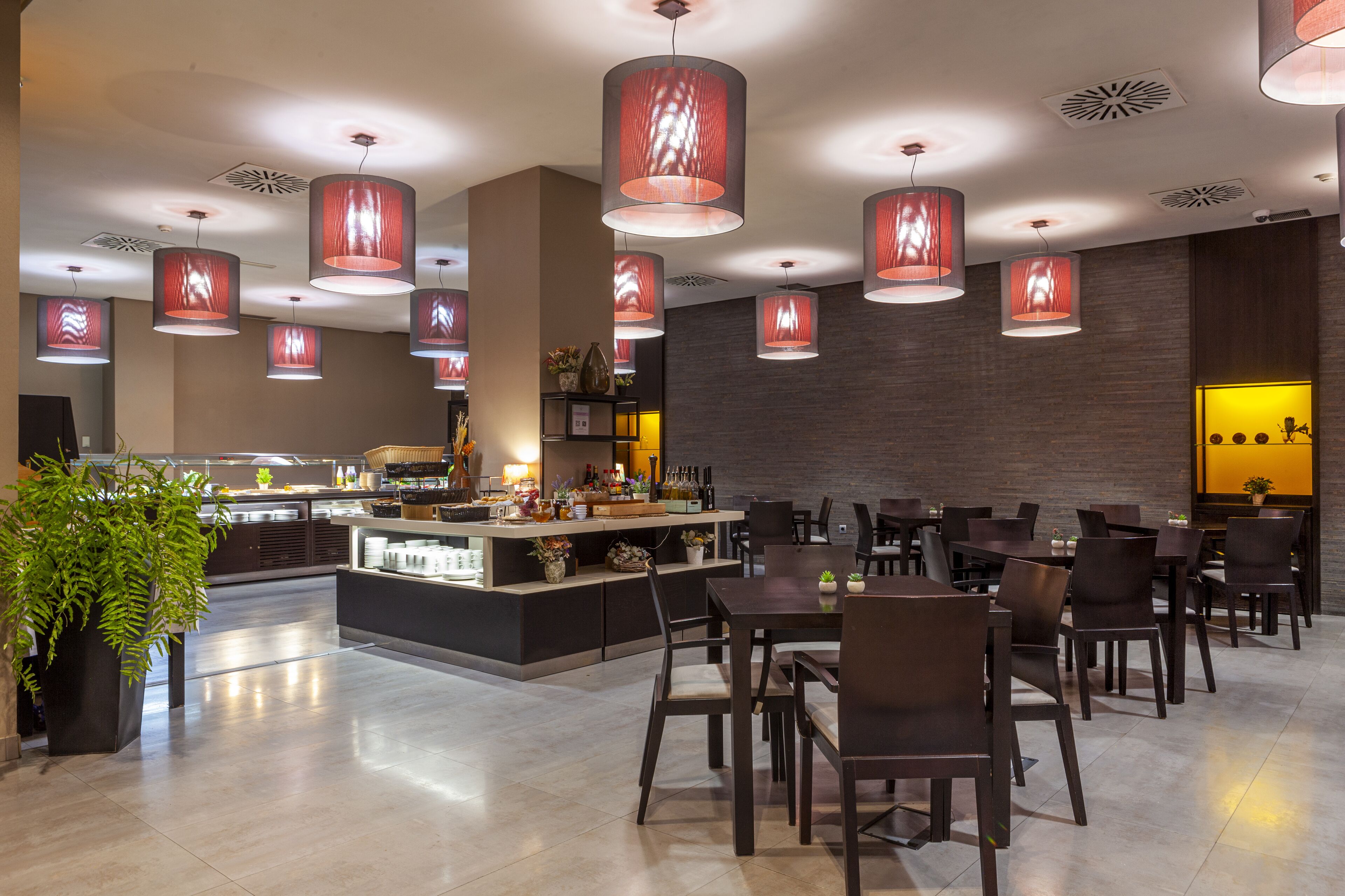 Ein modernes Restaurant mit einer zentralen Bar, mehreren Essplätzen und roten Hängeleuchten.