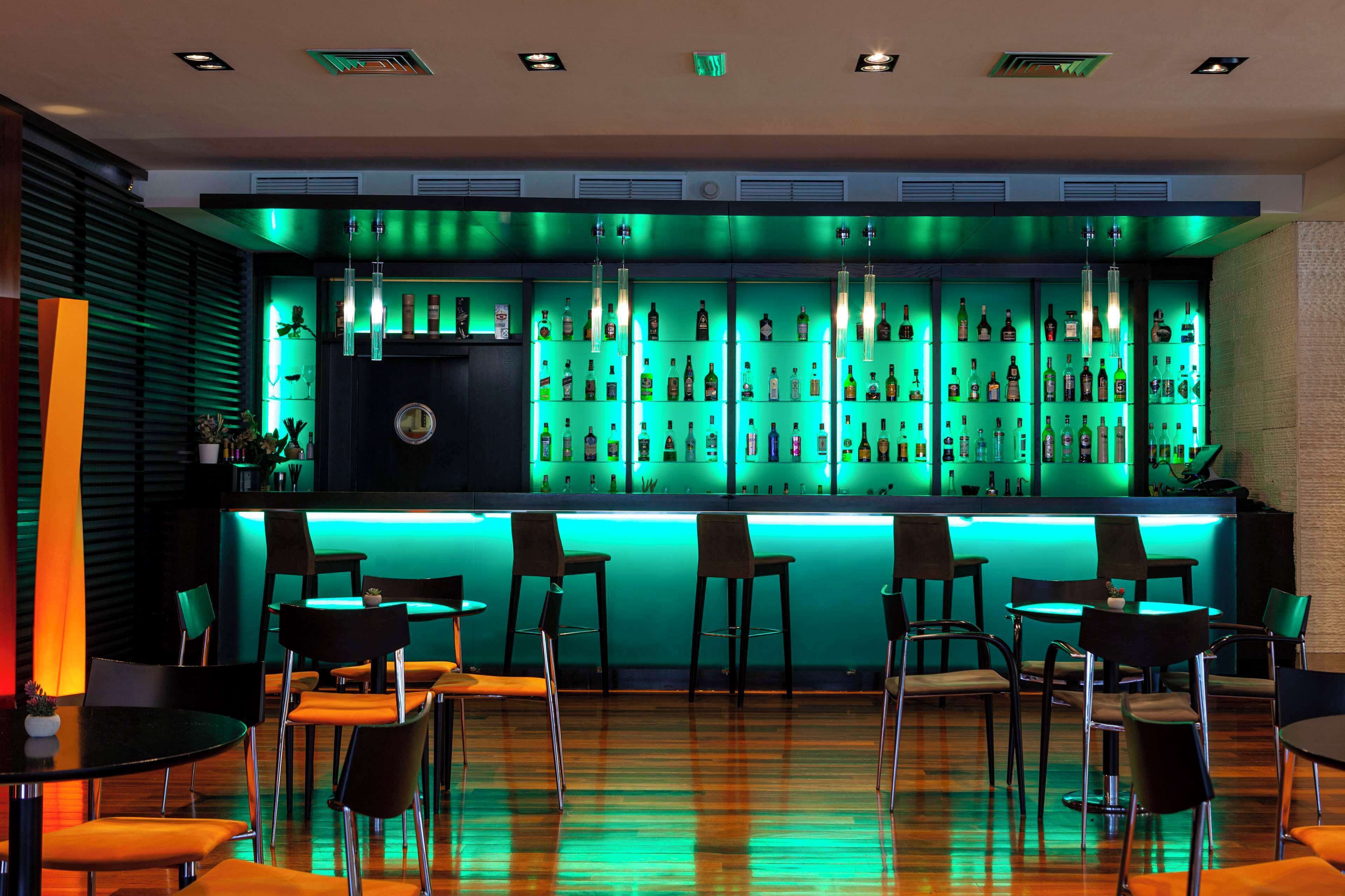 Zona de bar con mostrador iluminado en verde, varias sillas y una variedad de botellas en estanterías.