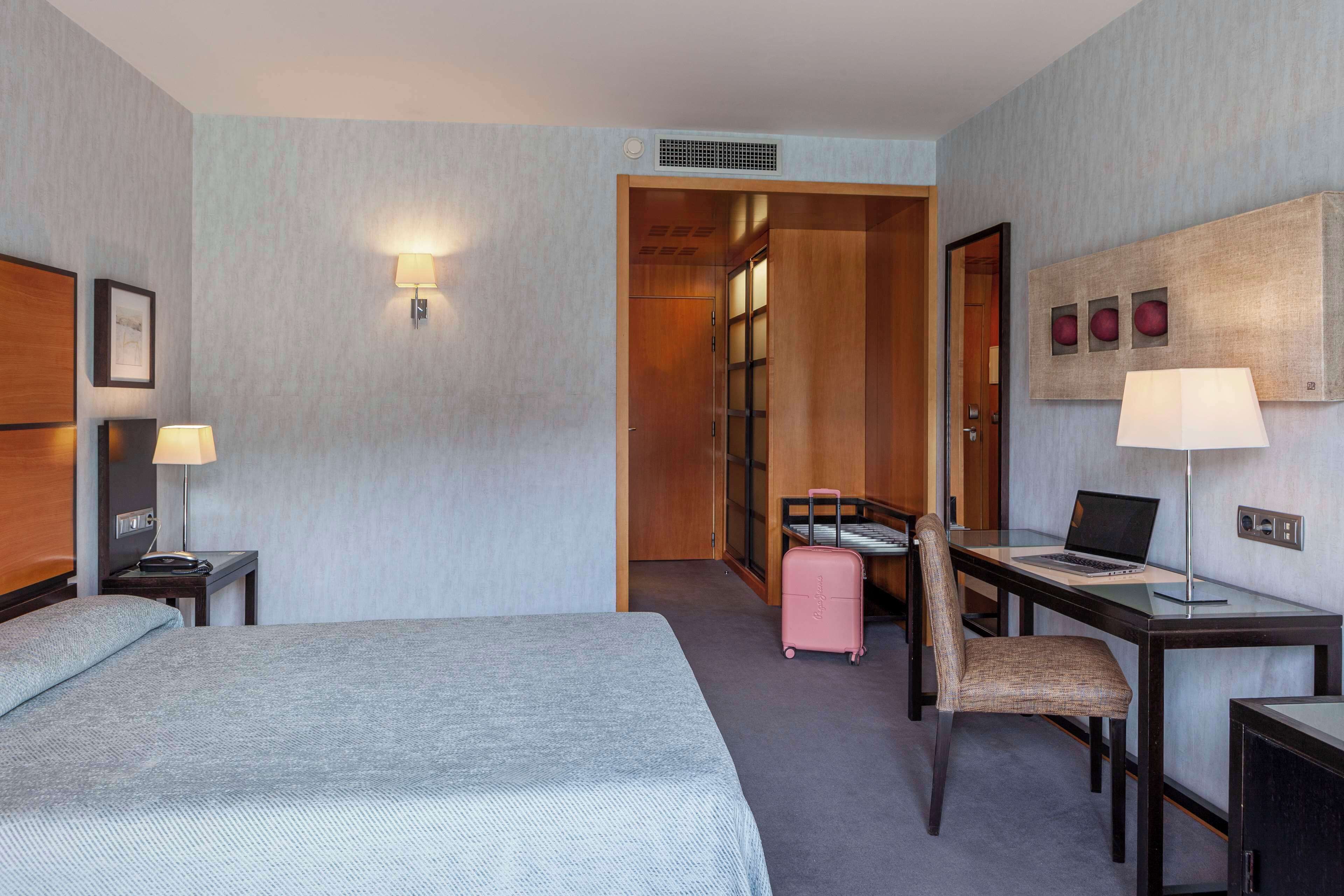 Habitación de hotel con una cama grande, un escritorio con una computadora portátil, una silla, una maleta y una lámpara fijada a la pared.