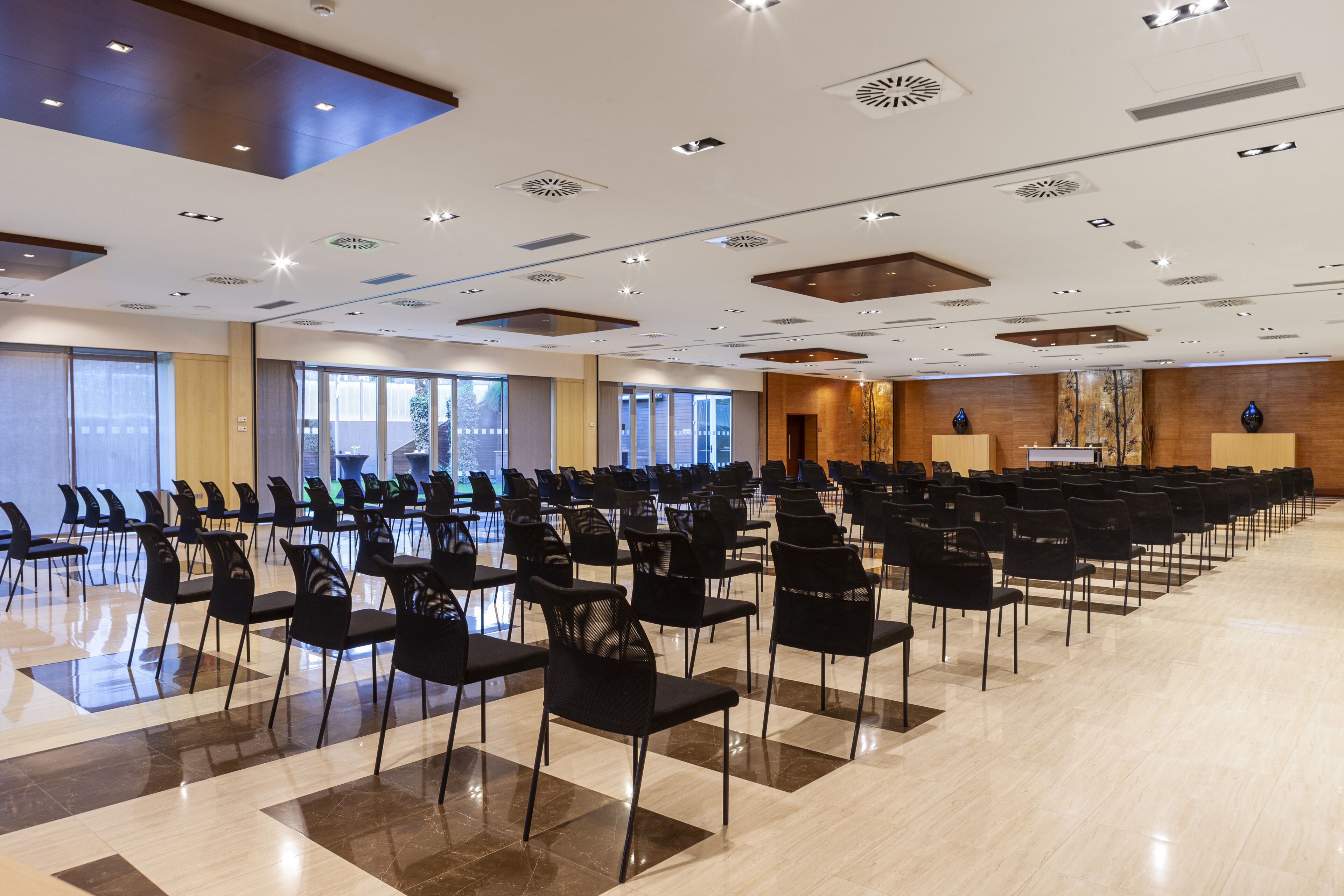 Une salle de conférence spacieuse, dotée de rangées de chaises noires, d’un sol en marbre poli et d’un plafond au design moderne avec des luminaires encastrés.