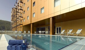 Zona de piscina de hotel con sillones de descanso y una mesa, barandilla de vidrio y vista a la montaña.