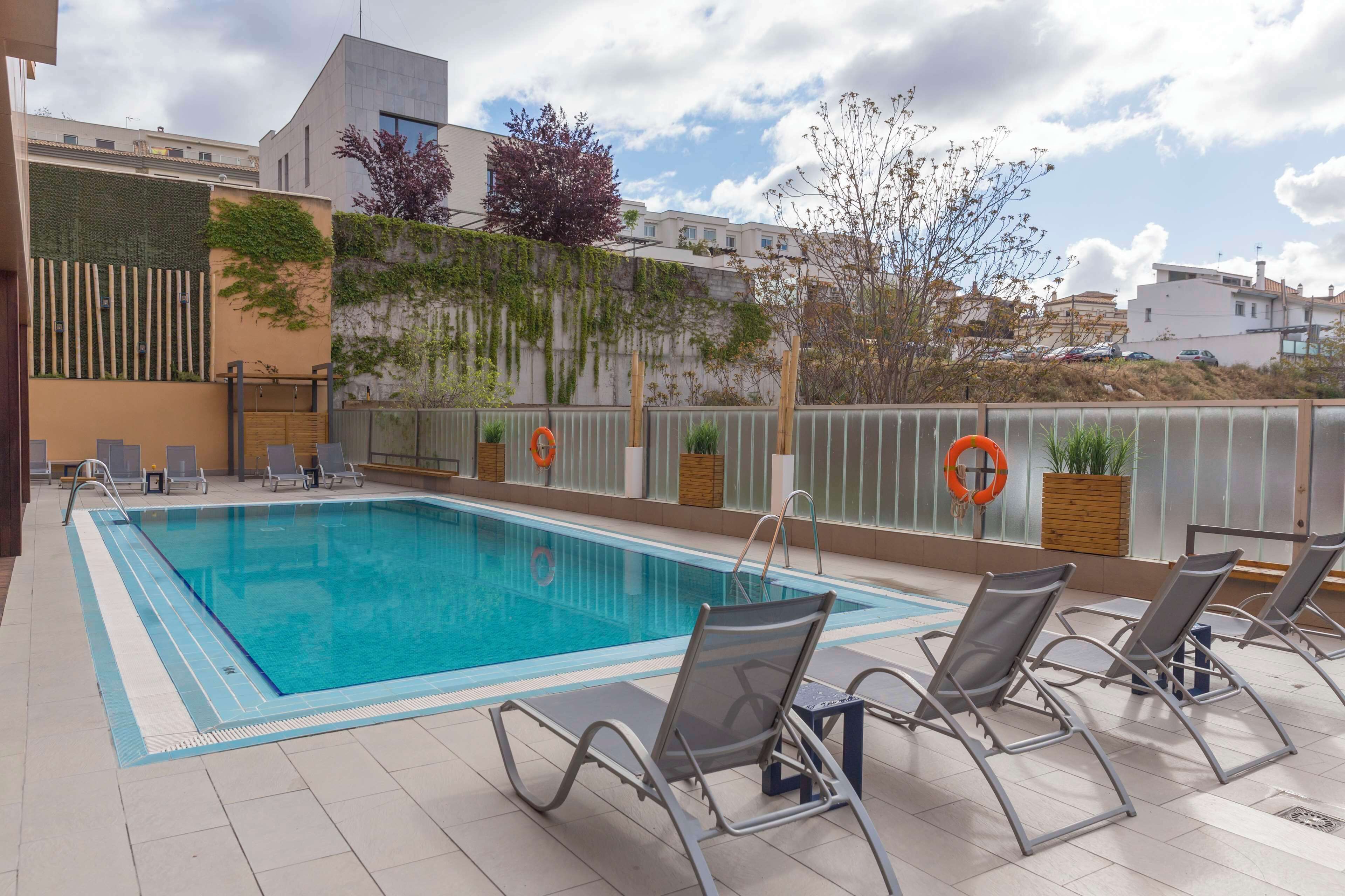 Une piscine avec des chaises longues, une bouée de sauvetage et une vue sur les bâtiments et les arbres.