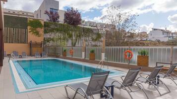 Une piscine avec des chaises longues, une bouée de sauvetage et une vue sur les bâtiments et les arbres.