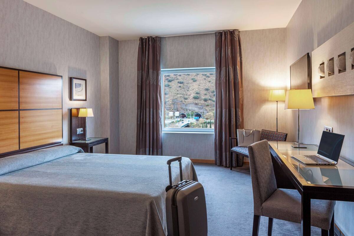 Habitación de hotel con una cama, un escritorio con una computadora portátil, una silla, una maleta y una ventana con cortinas.