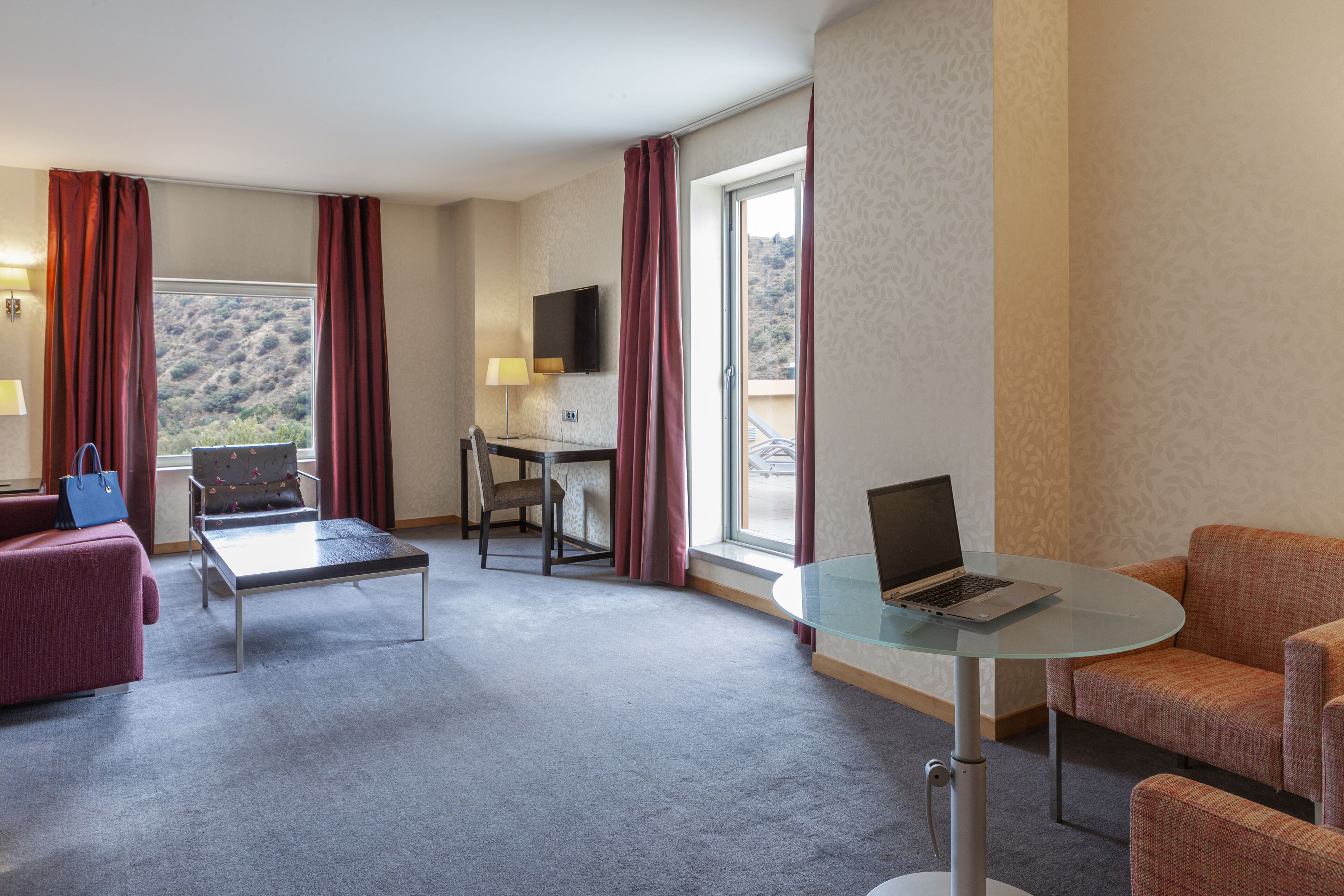 Quarto de hotel com mesa de vidro, laptop, sofá, poltrona e televisão.