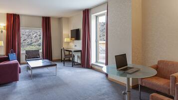 Quarto de hotel com mesa de vidro, laptop, sofá, poltrona e televisão.