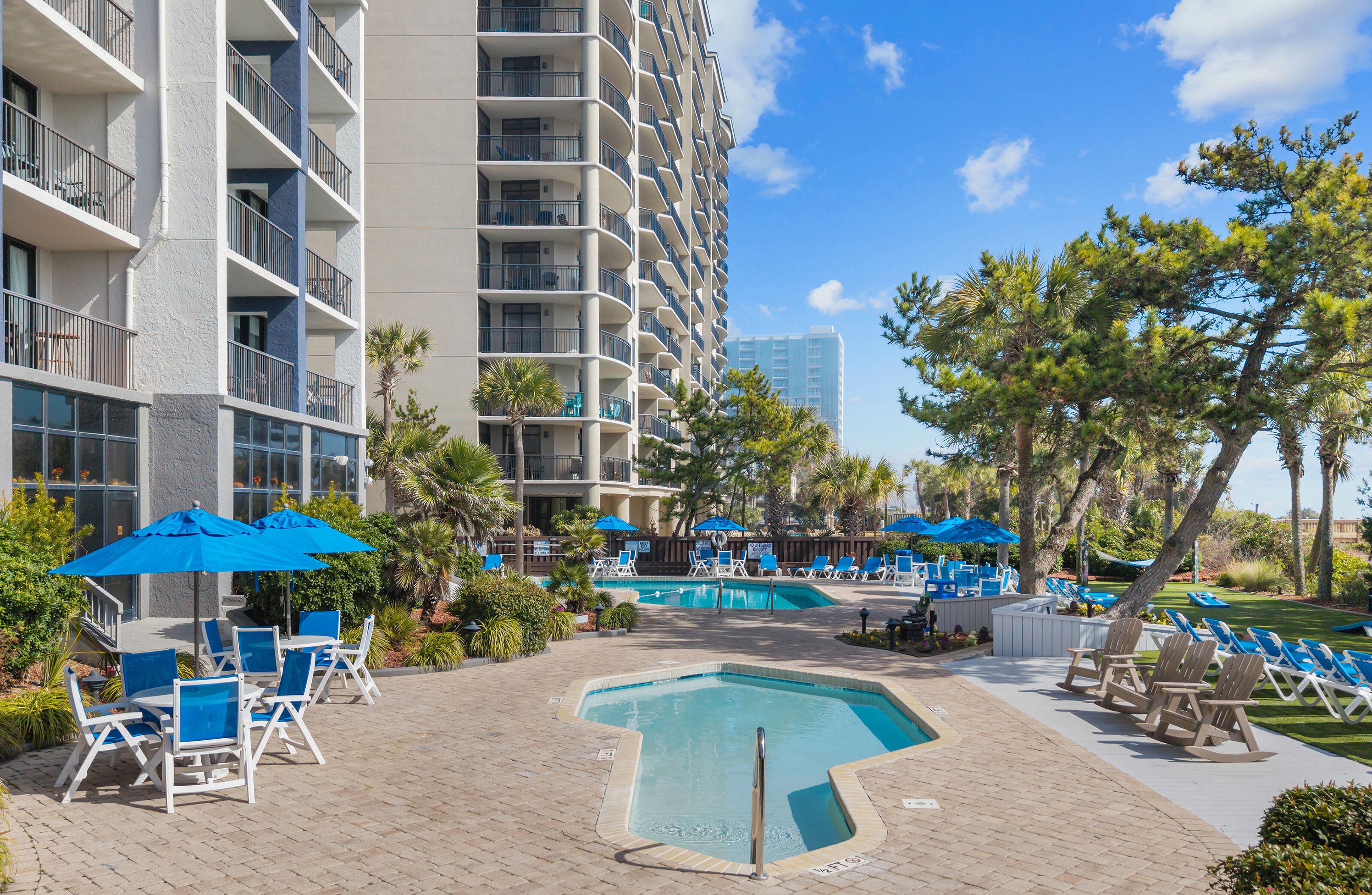 A hotel pool area with lounge chairs, umbrellas, and a large building in the background.