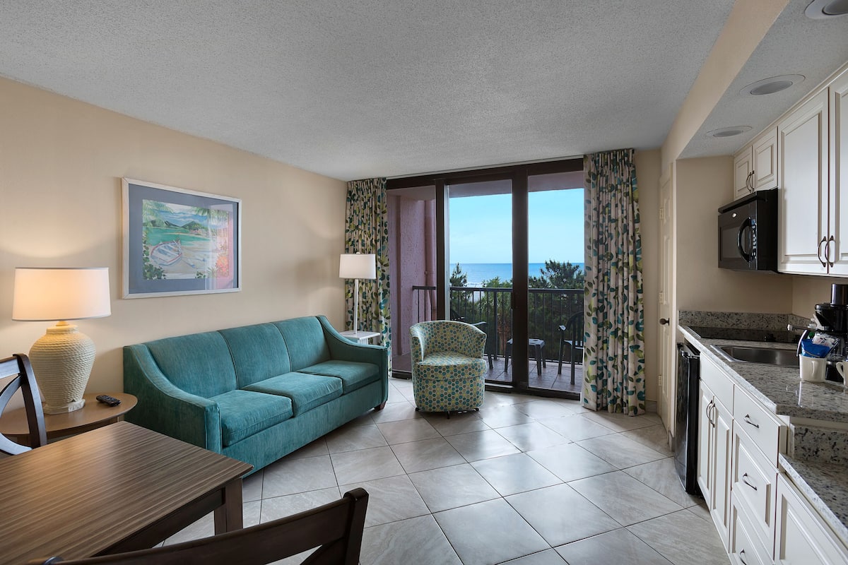 A hotel room with a teal sofa, a patterned armchair, a dining table, a kitchen area, and a large window with a view of the sea.