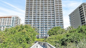 Un edificio de gran altura con varios balcones, rodeado de abundante vegetación y un puente peatonal.