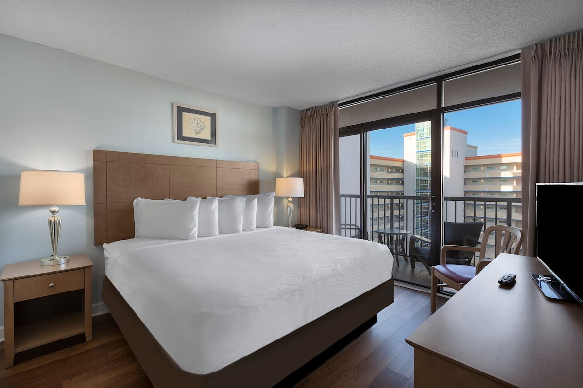 A hotel room with a large bed, two bedside tables with lamps, a desk with a TV, and a balcony with a view of buildings.