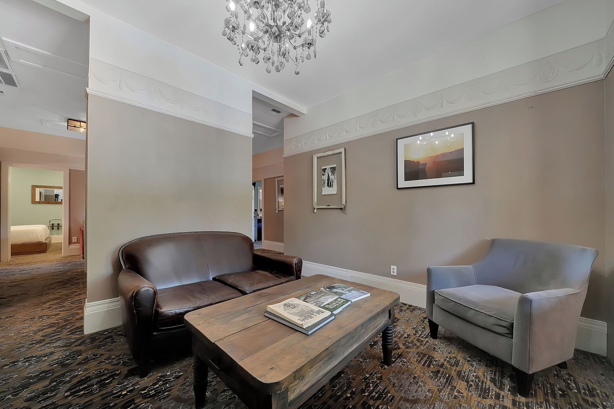A hotel room with a brown leather sofa, a grey armchair, a wooden coffee table with magazines, and a chandelier.
