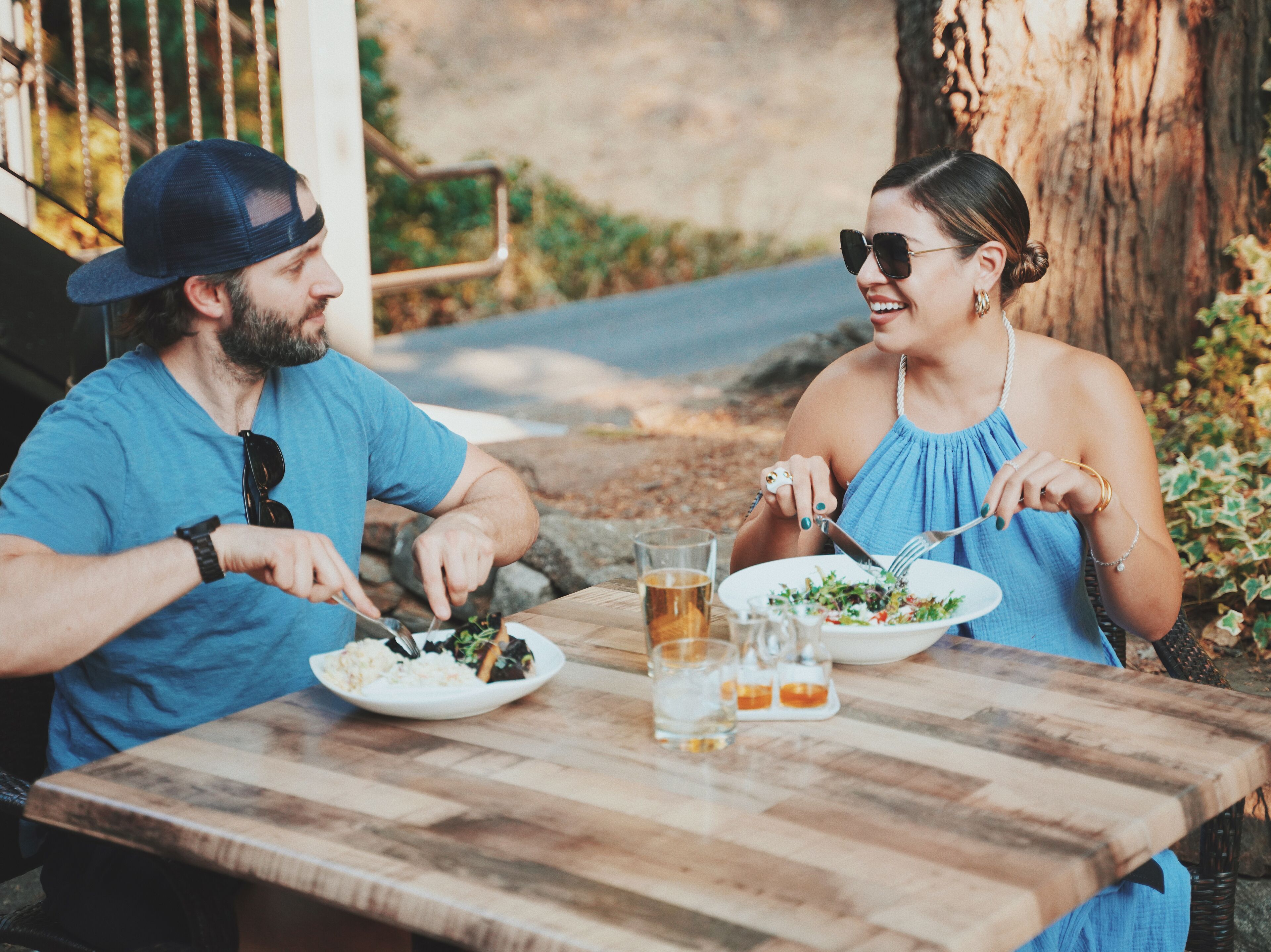 Two people dining at an outdoor table with a wooden surface, surrounded by greenery and a tree trunk.