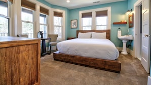 A bedroom with a wooden bed, a desk, and a chair. There are windows with blinds, a sink, and a mirror. - The Groveland Hotel (Groveland)