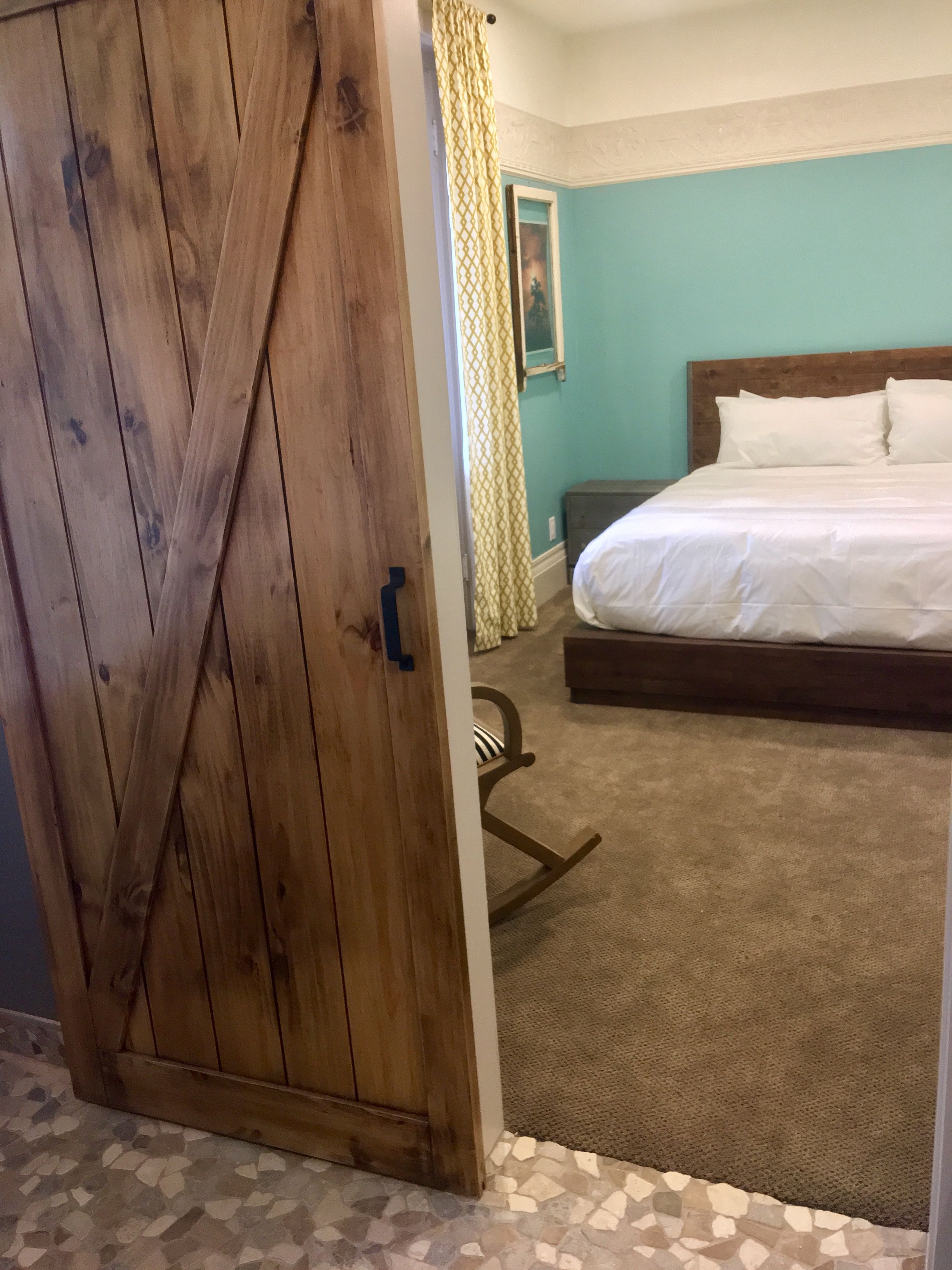Ein Schlafzimmer mit Holztür, einem Bett mit weißer Bettwäsche, einem Schaukelstuhl und einem gemusterten Vorhang.