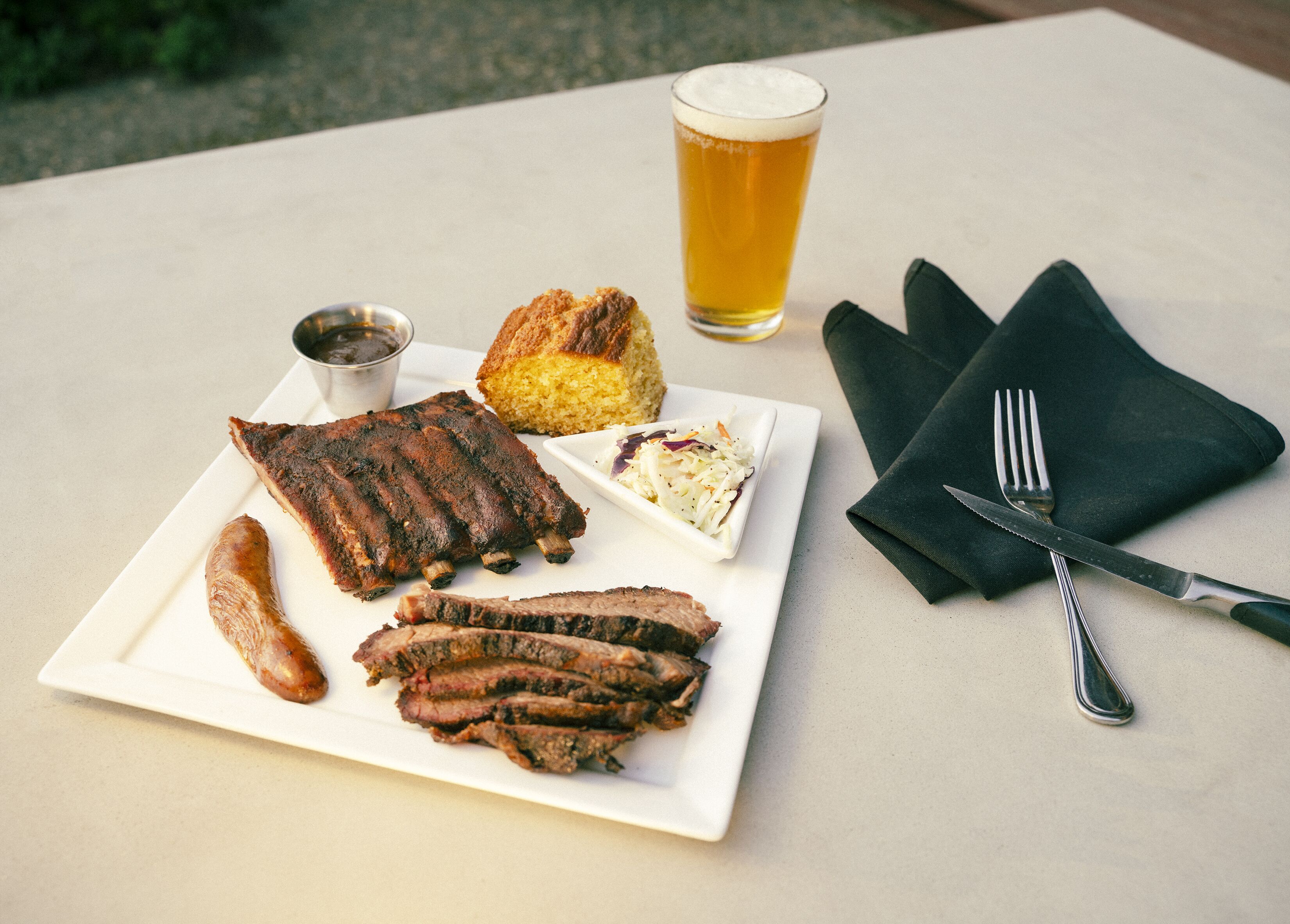 Un plato de costillas, un bollo, ensalada de col y una salchicha sobre un plato blanco, una jarra de cerveza y un tenedor sobre una servilleta en una mesa.