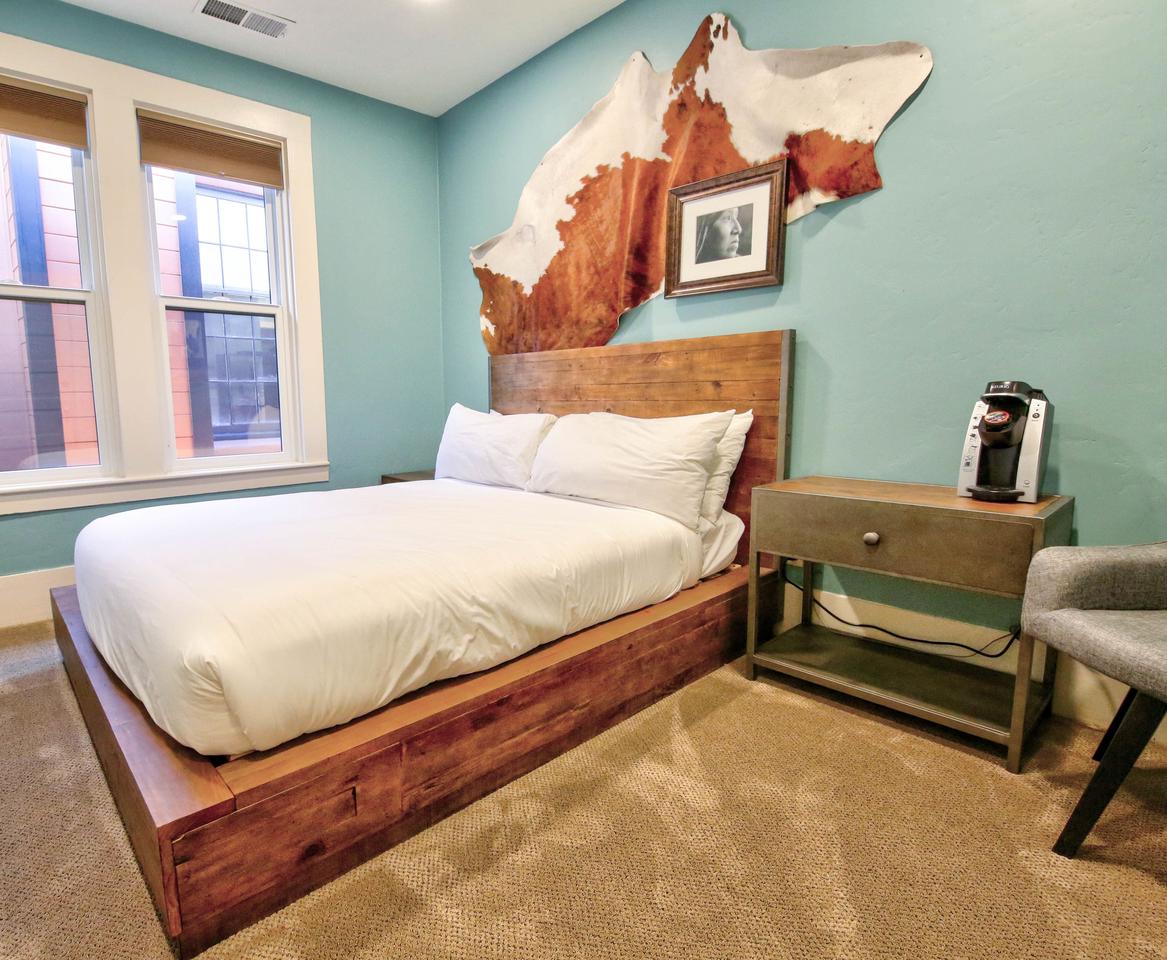 A bedroom with a wooden bed, a nightstand with a coffee maker, a framed picture, and a large animal hide on the wall.
