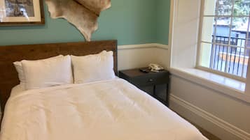 Una cama bien tendida con sábanas blancas, cabecera de madera, un cuadro enmarcado en la pared, una mesita de noche con teléfono y control remoto, y una ventana con vistas al exterior.