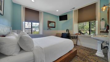 A bedroom with a large bed, a bedside table, a TV, a window with blinds, and a bathroom with a sink and mirror.