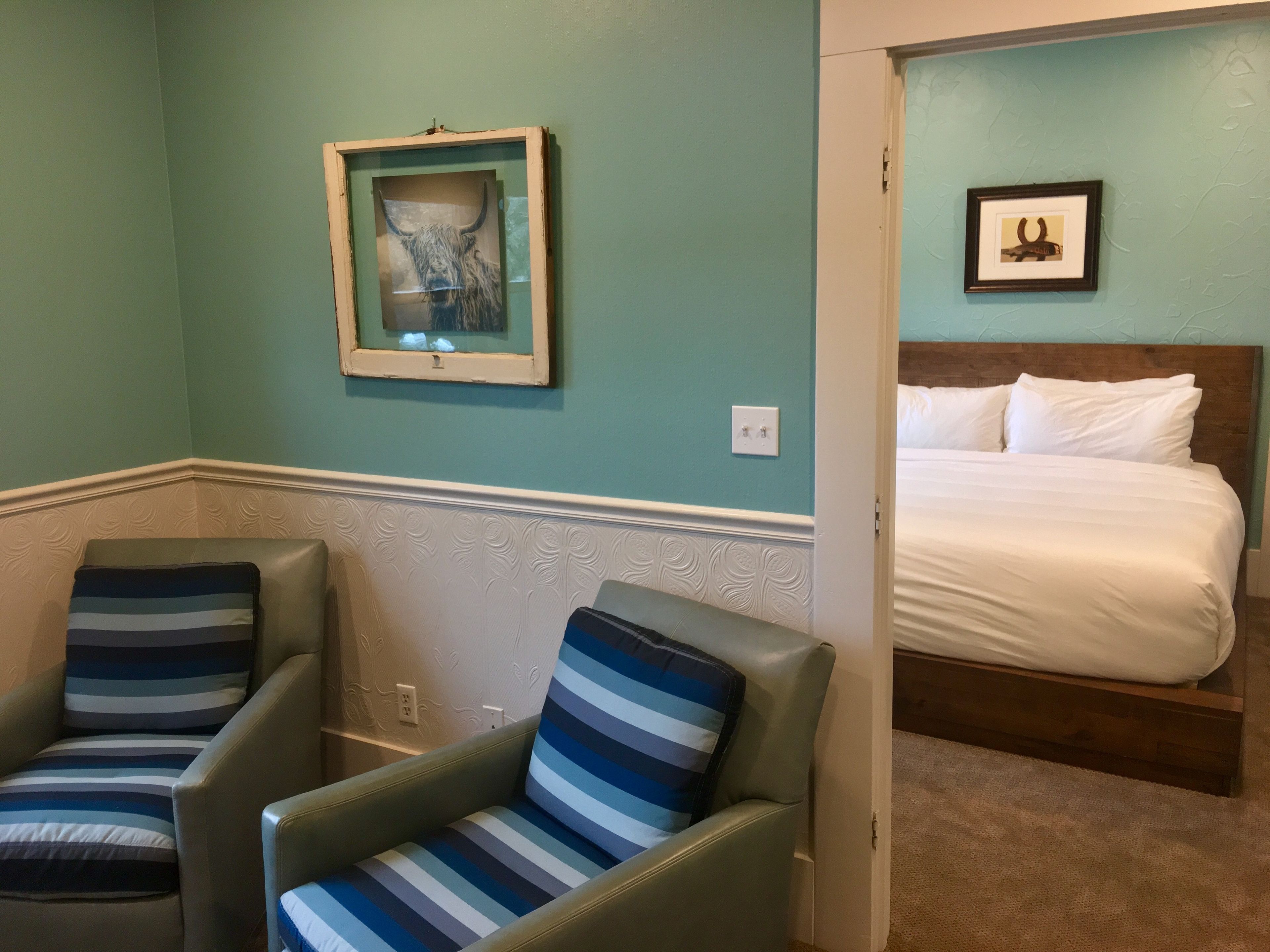 Une pièce aux murs de couleur turquoise, un lit recouvert d’une literie blanche, deux fauteuils à rayures et, au mur, un tableau représentant un taureau.