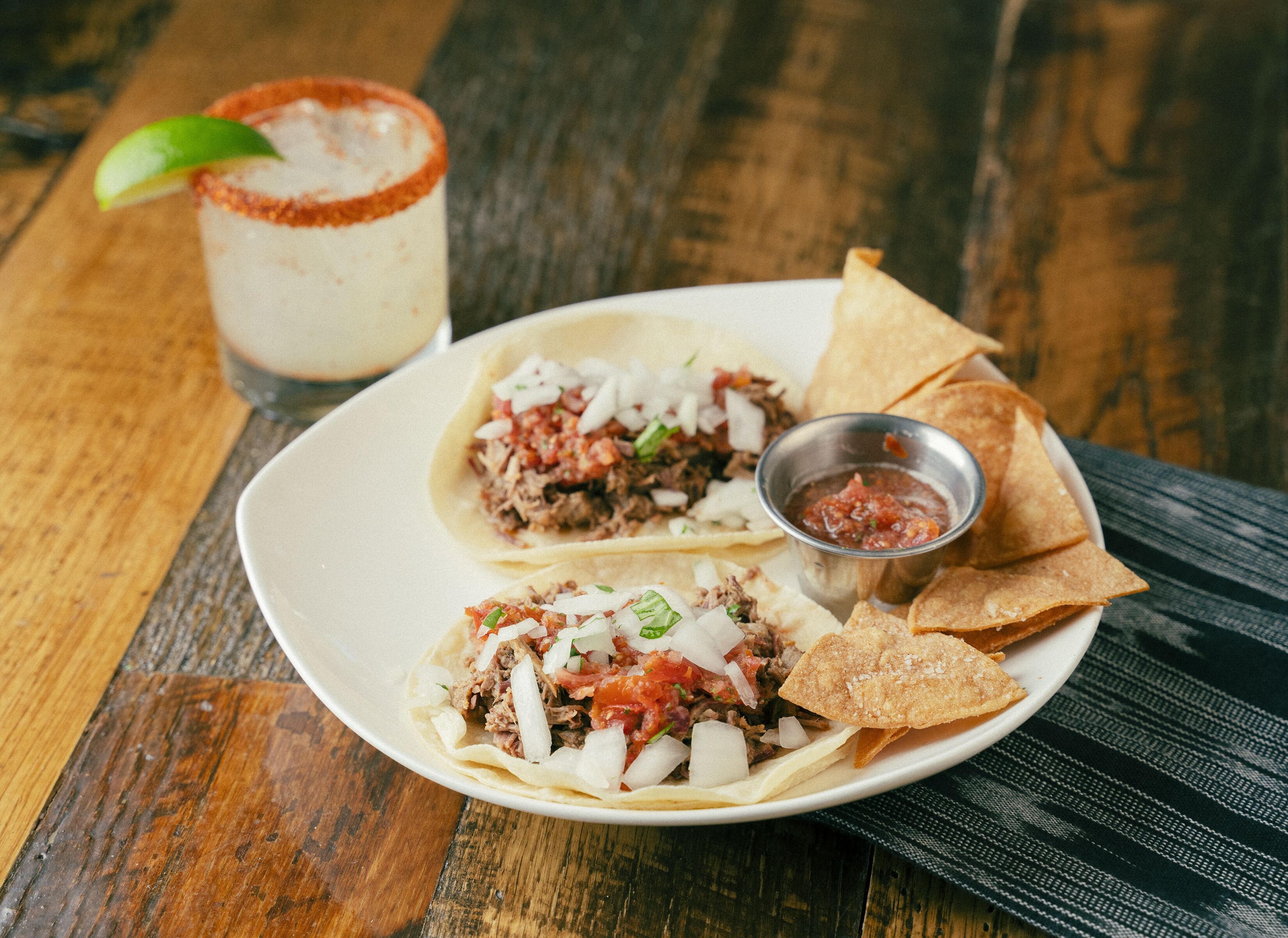 A plate of tacos with a side of chips and salsa, and a margarita on a wooden table.