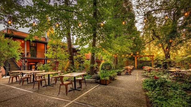 An outdoor patio area with tables and chairs, surrounded by trees and plants, and string lights hanging overhead. - The Groveland Hotel (Groveland)