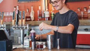 A bartender preparing a drink at a bar with various bottles and a chalkboard menu in the background. - The Groveland Hotel (Groveland)