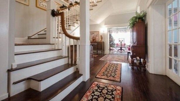 Un magnifique escalier en bois foncé, avec des marches et des rampes blanches, mène à un couloir bien éclairé, agrémenté de tapis à motifs et d’une grande armoire en bois.