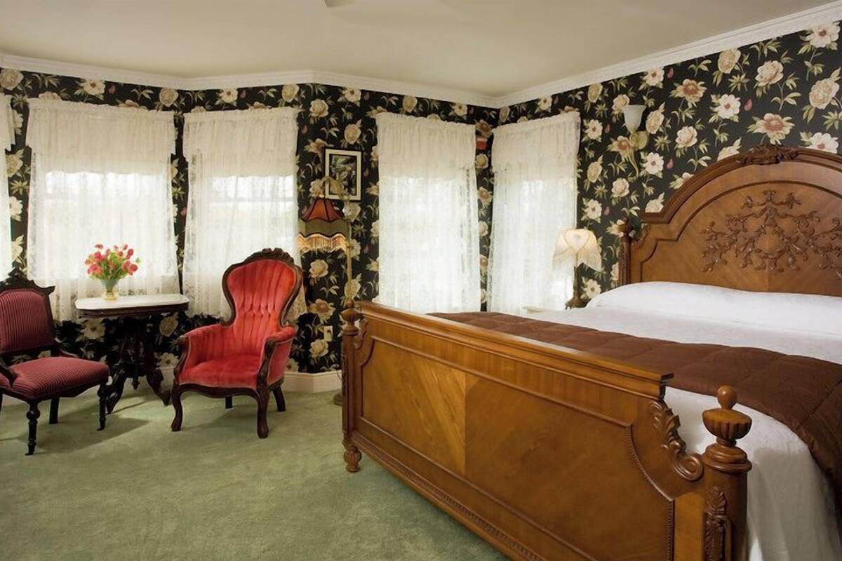 A bedroom with a wooden bed, a red chair, a small table with a vase of flowers, and floral wallpaper.