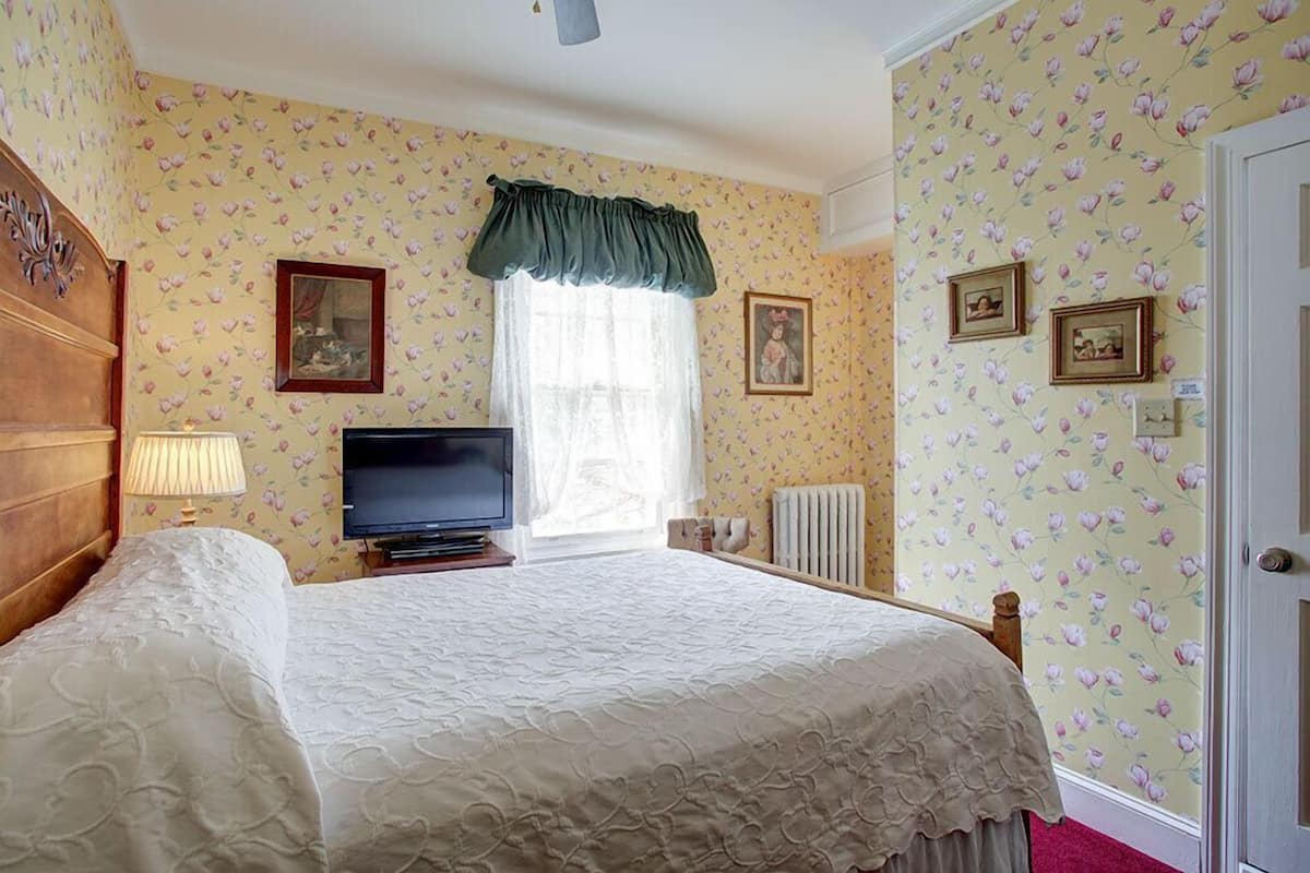 A bedroom with floral wallpaper, a bed with a white bedspread, a television on a stand, and a window with a green valance.