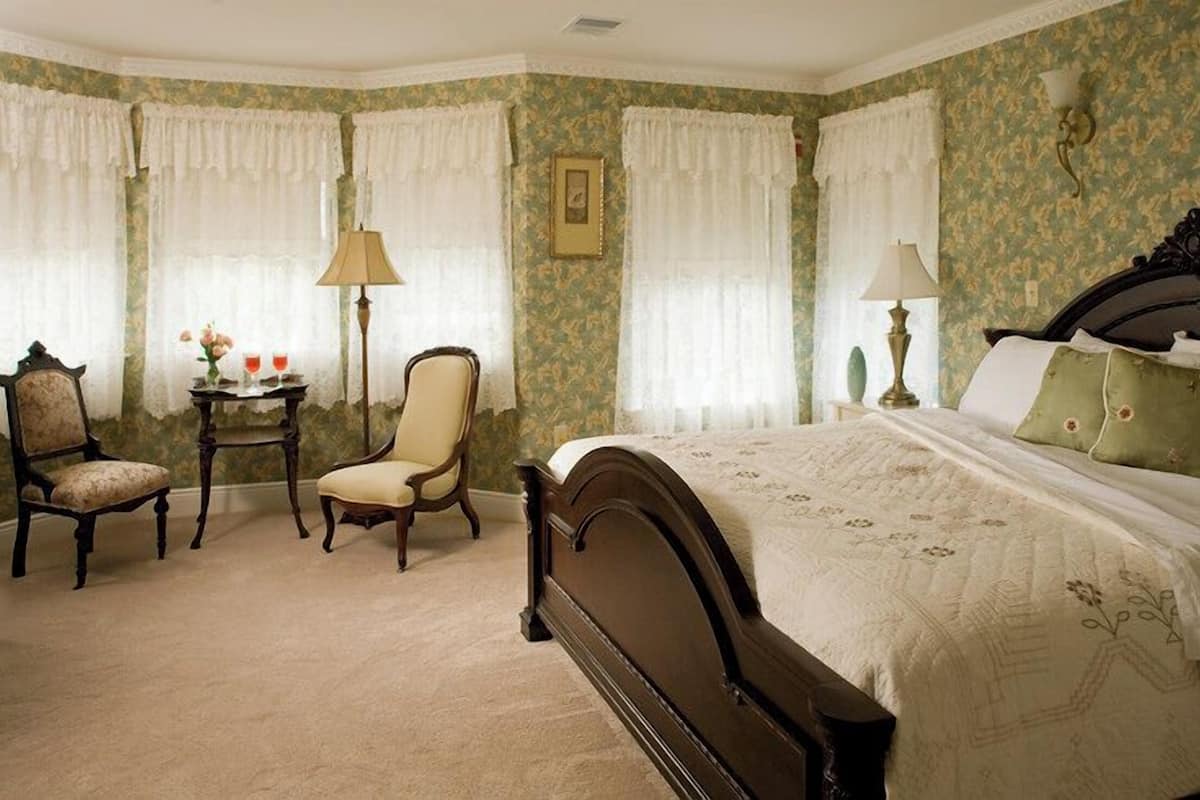 A bedroom with a large bed, two chairs, a small table with a lamp, and a picture on the wall.