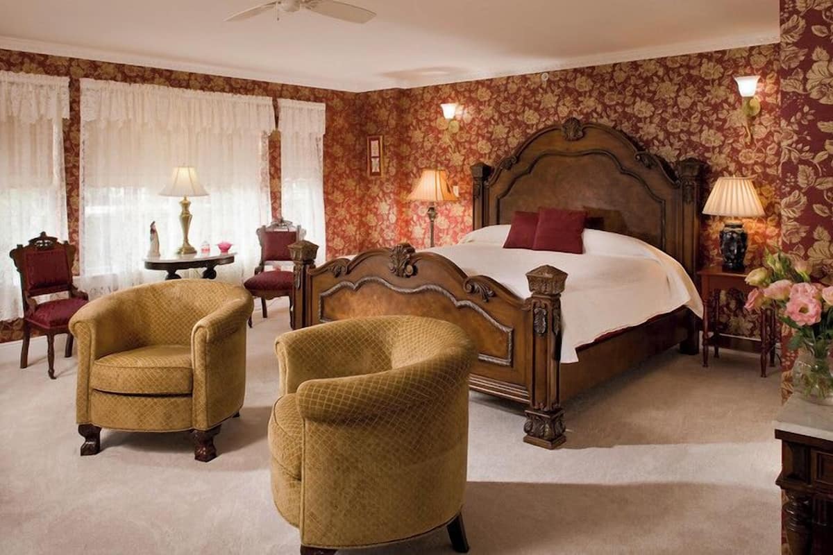 A classic bedroom with a four-poster bed, two armchairs, a nightstand with a lamp, and floral wallpaper.