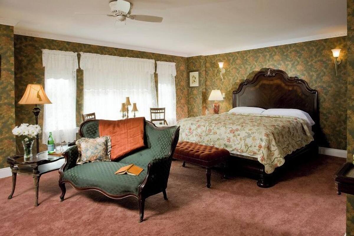 A traditional bedroom with a bed, a green armchair, a wooden nightstand, and a table with a bowl of fruit.