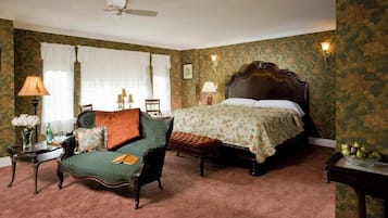 Quarto tradicional com uma cama, uma poltrona verde, uma mesinha de cabeceira de madeira e uma mesa com uma tigela de frutas.