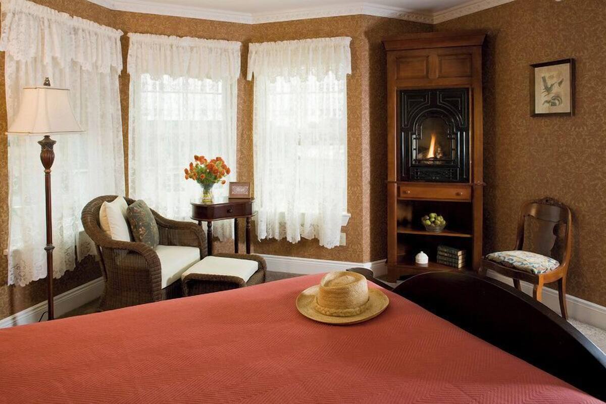 A room with a fireplace, a bed with a red bedspread, two chairs, a table with a vase of flowers, and a lamp.