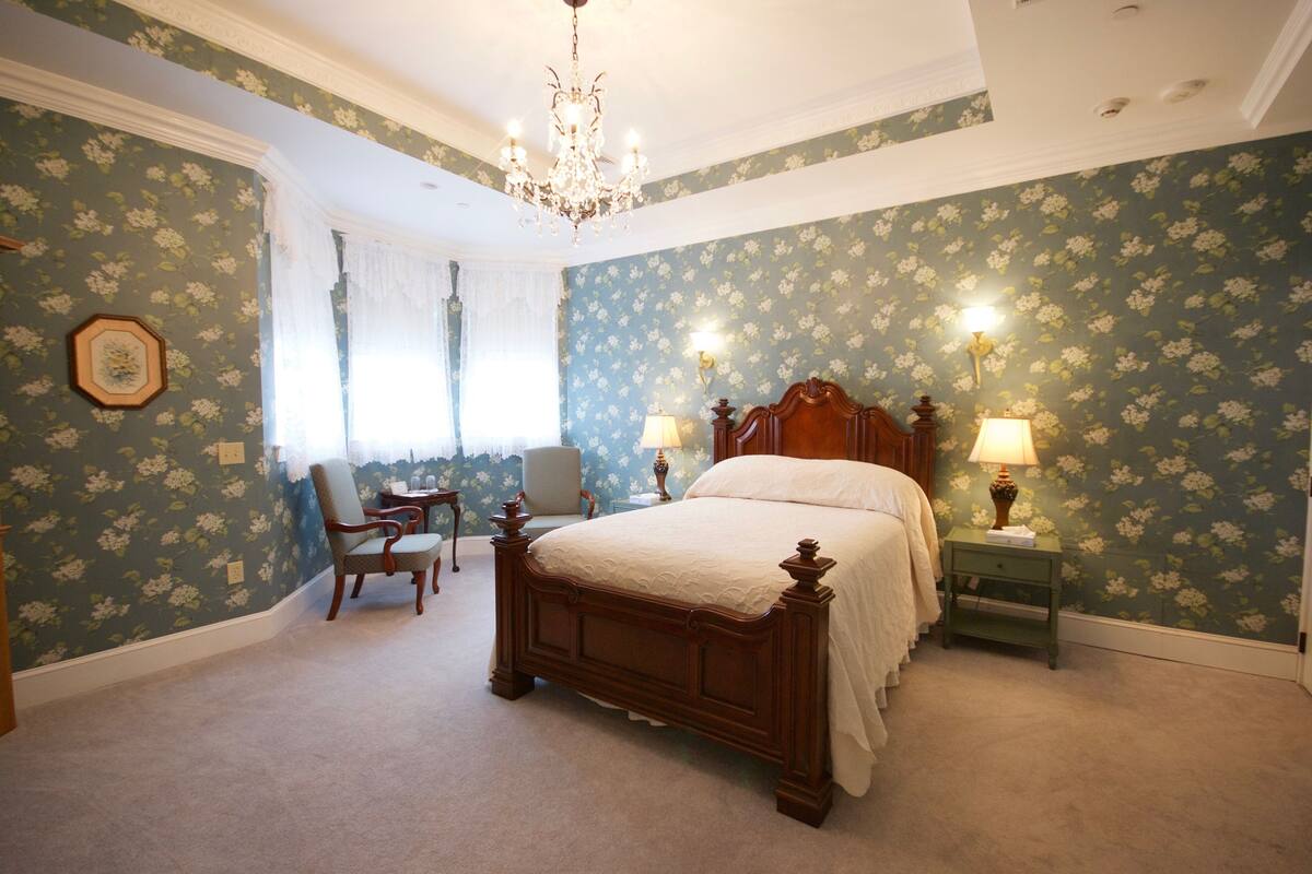 A bedroom with floral wallpaper, a wooden bed, two bedside lamps, a chair, and a chandelier.