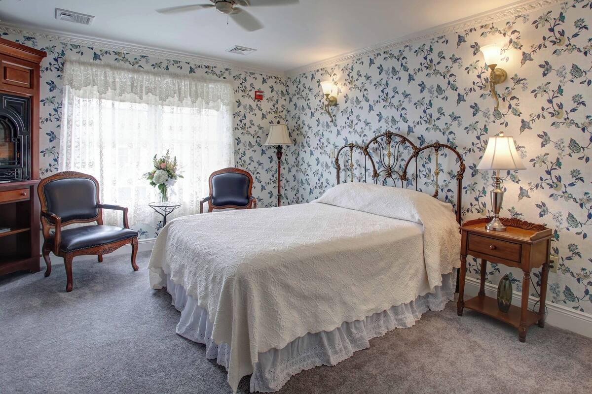 A bedroom with a bed, two chairs, a nightstand with a lamp, a ceiling fan, floral wallpaper, and a window with curtains.